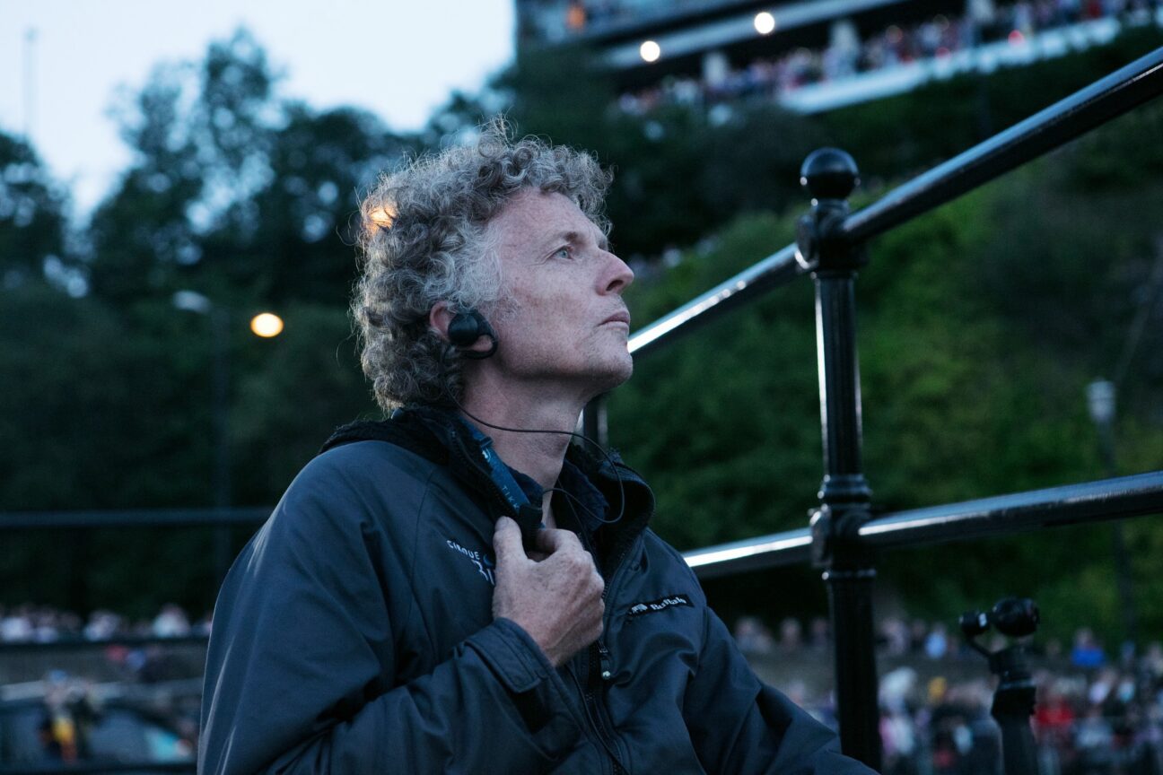 Billy Alwen looking intently upwards to the right of the screen, he has an ear piece in.