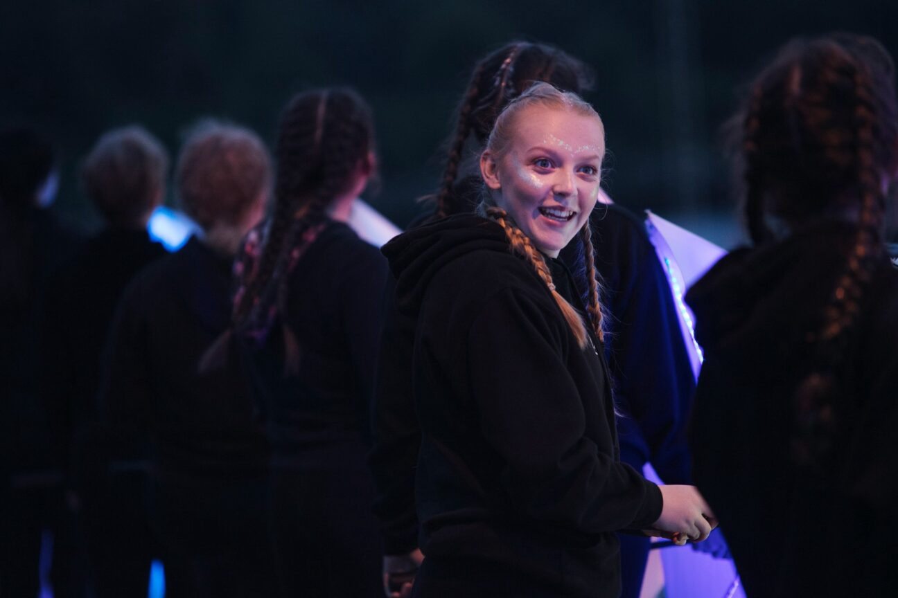 A teenage girl faces out with glitter on her cheeks and forehead smiling past the camera. She is wearing black and is in a line of other young people also wearing black, they are facing the front.