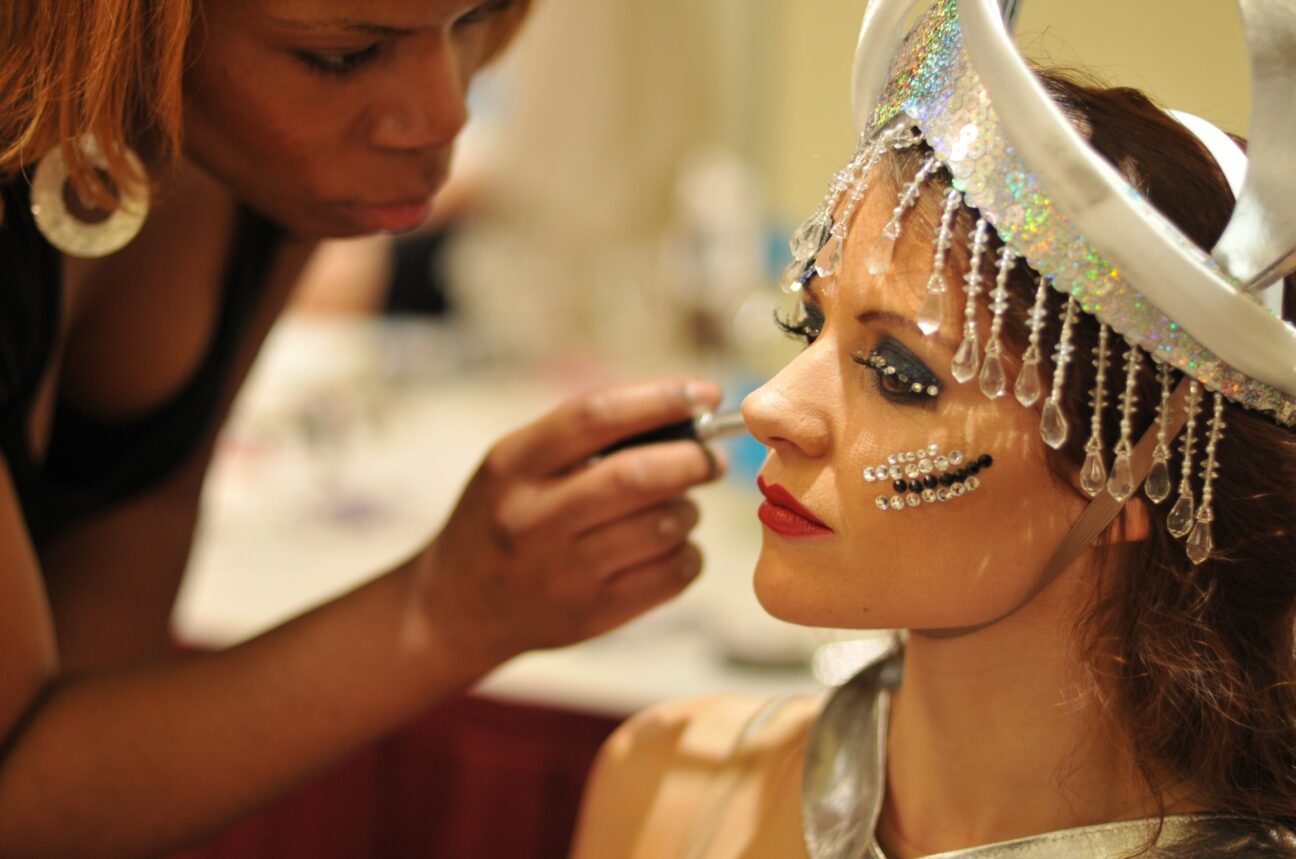 Performer having her makeup done. She has silver and black gems on her face and a sparkly headdress.