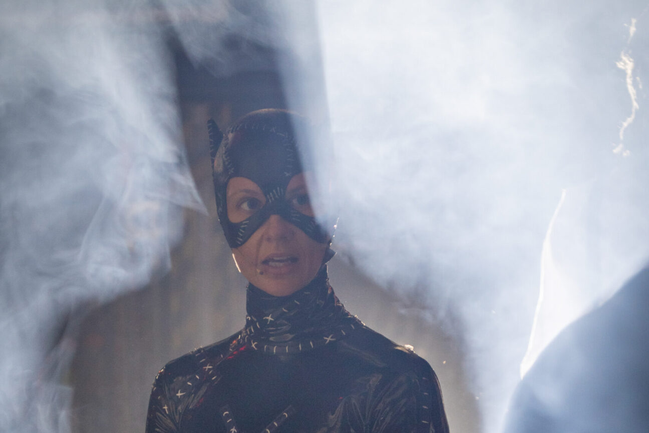 A female performer in a head mic, wearing a black batman costume surrounded by smoke.