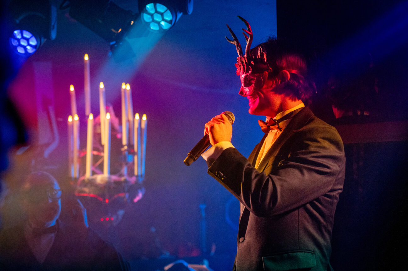 A man stands, in suit and bowtie, with a microphone and a mask with antlers. He is smiling and lit softly by a red light as well as light from a candelabra displayed behind him to his left.