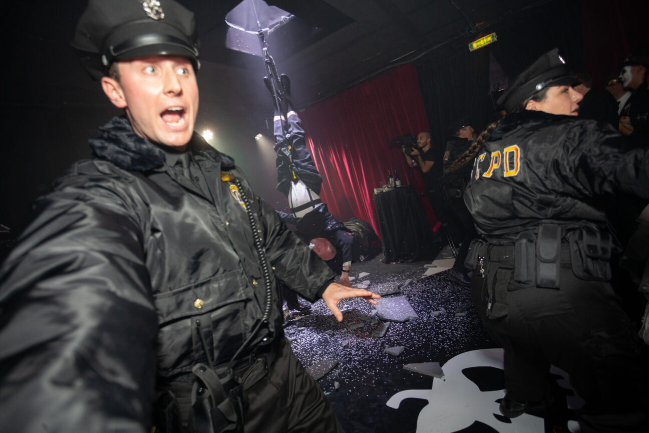 2 performers dressed as Police officers usher audience away from a "crime scene" where a man is suspended from the ceiling by his feet. There is debris on the floor.