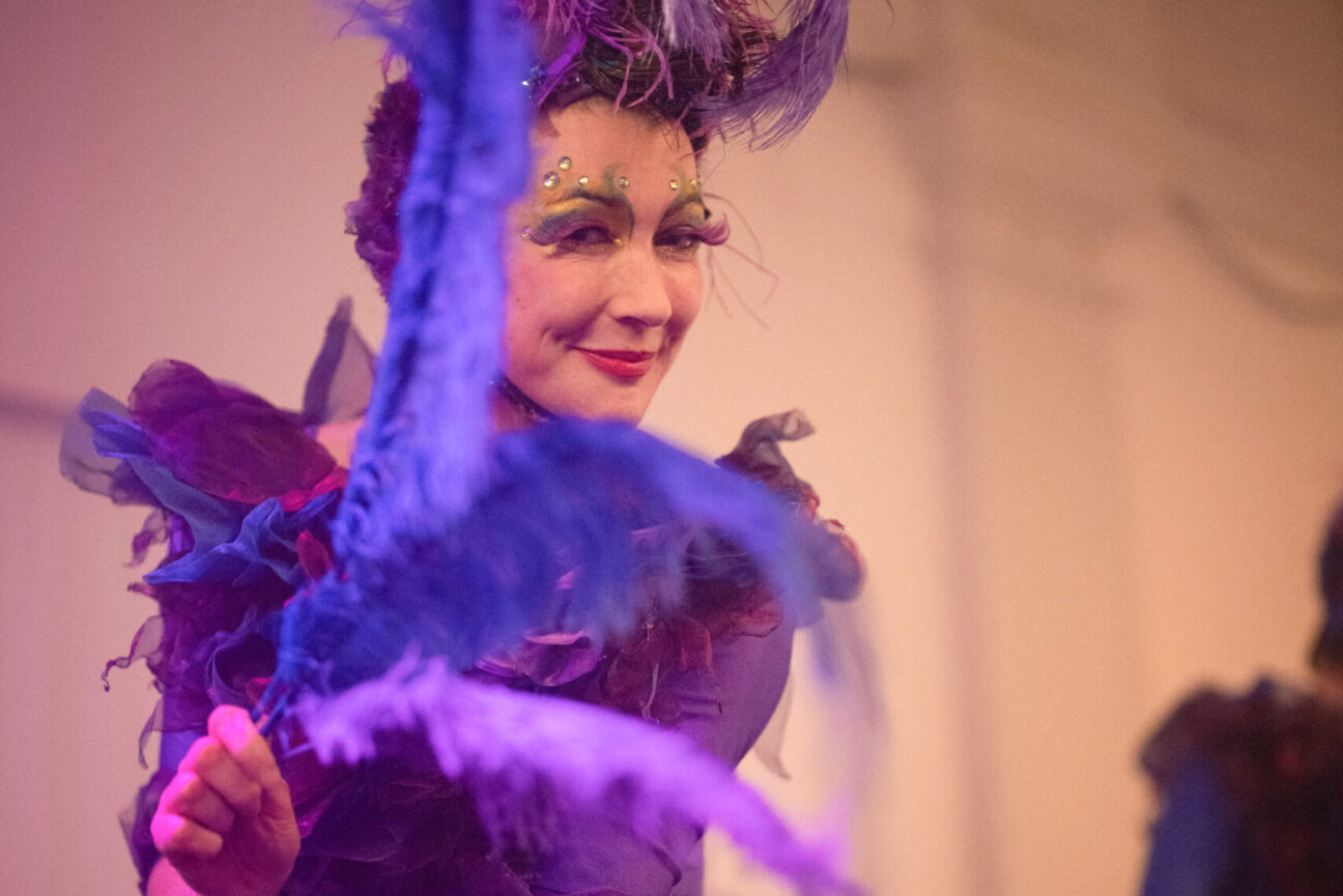 A female performer wears eccentric makeup with purple eyelashes and silver gems. She is waving large blue feather towards the camera whilst smiling. She is wearing a frilly and feathery purple outfit and headdress.