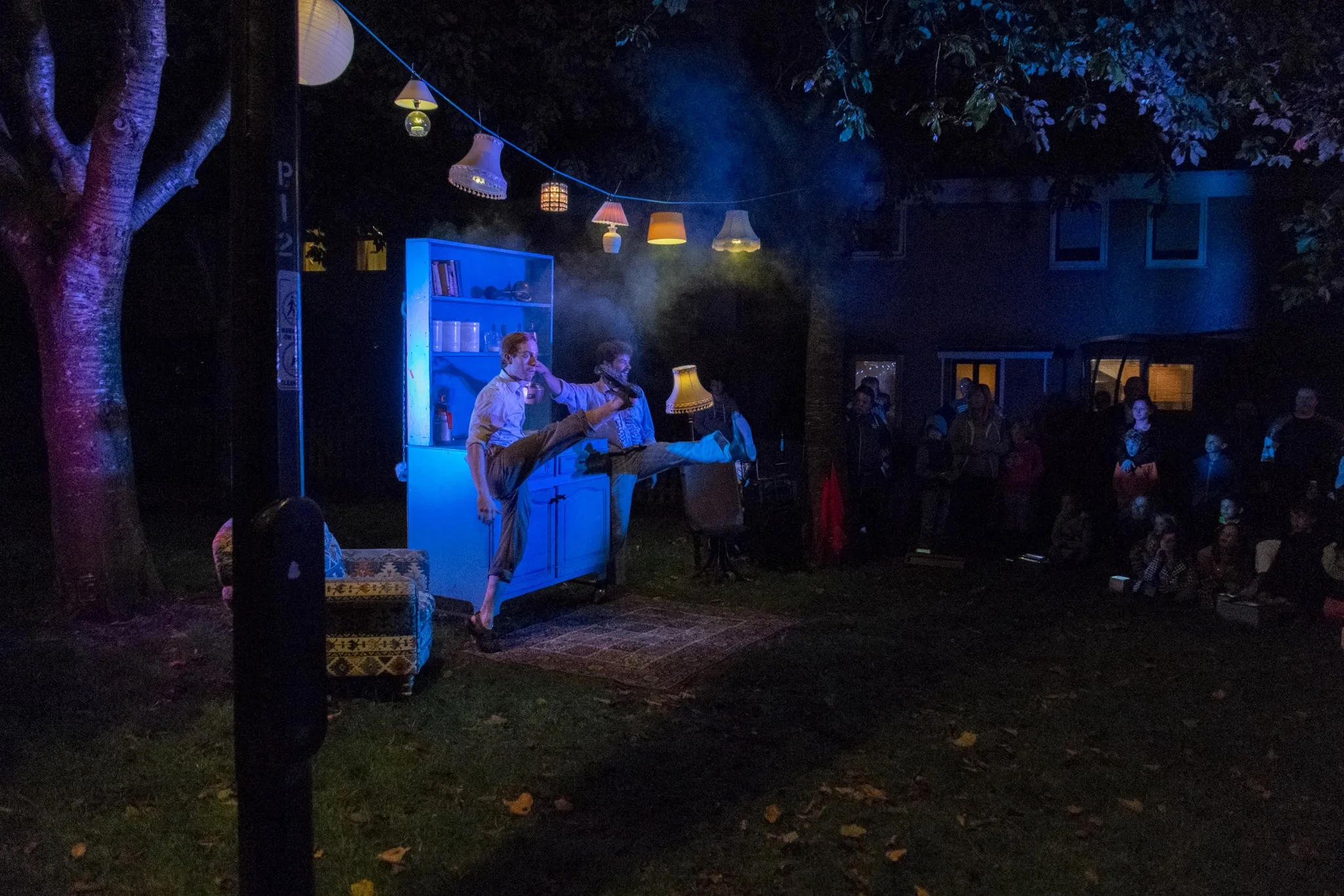 Kitchen Circus Project, 2015. Two performers are stood infant of a blue cupboard kicking their outside leg forwards towards where the audience are stood. Above them is a washing line of various lampshades. There is a rug on a grass floor, the performance is outside, there is also an armchair to the side.