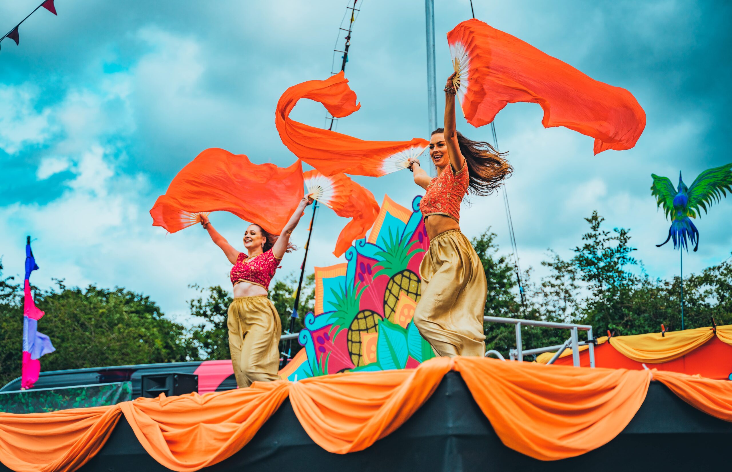 Party on the Green, two female performers wave orange fan pots in front of Cirque Bijou backdrops.