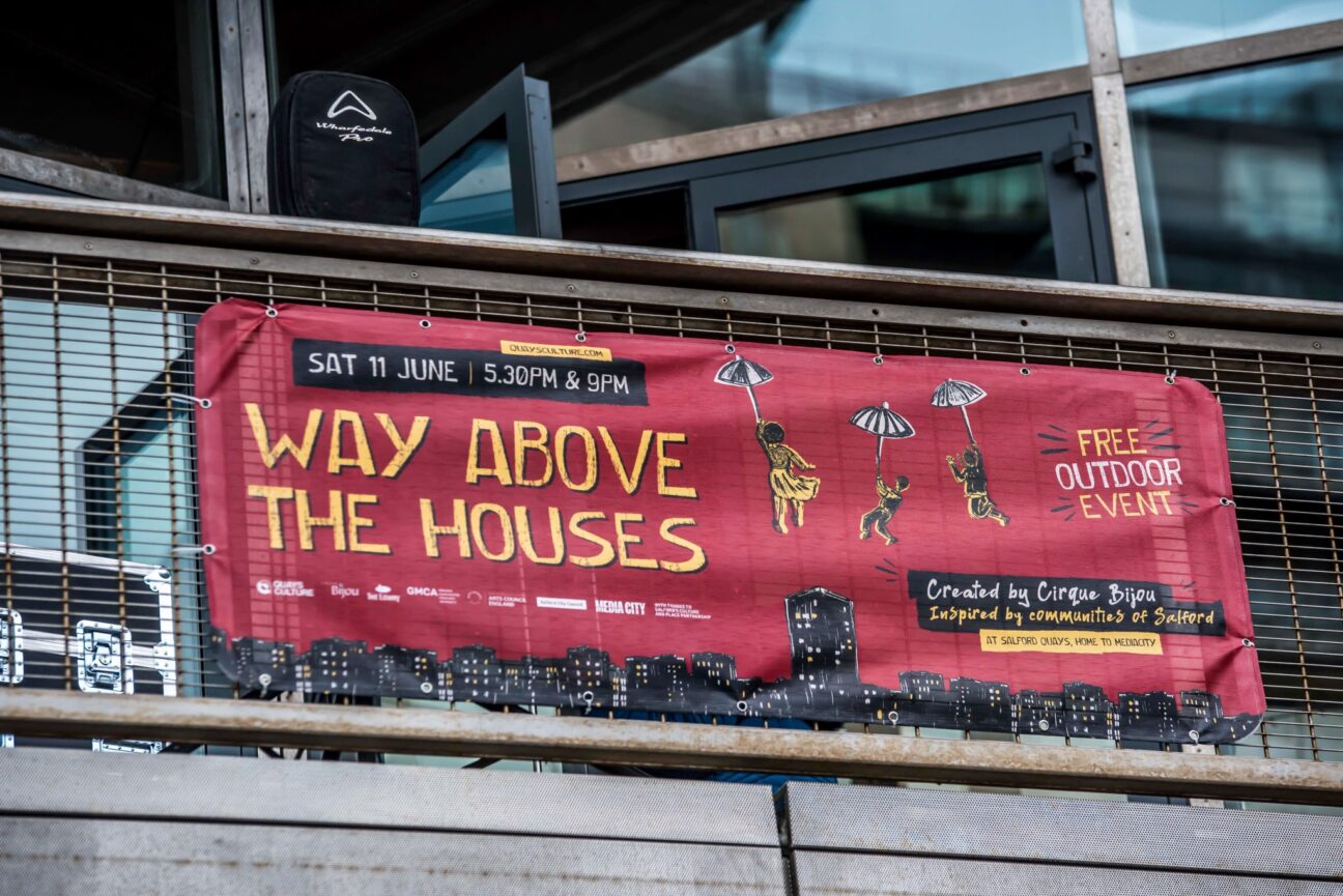 A red and yellow banner advertising the Way Above the Houses "Free Outdoor Event", created by Cirque Bijou and Inspired by communities of Salford. The date was Saturday June 11th, 5:30pm & 9pm.