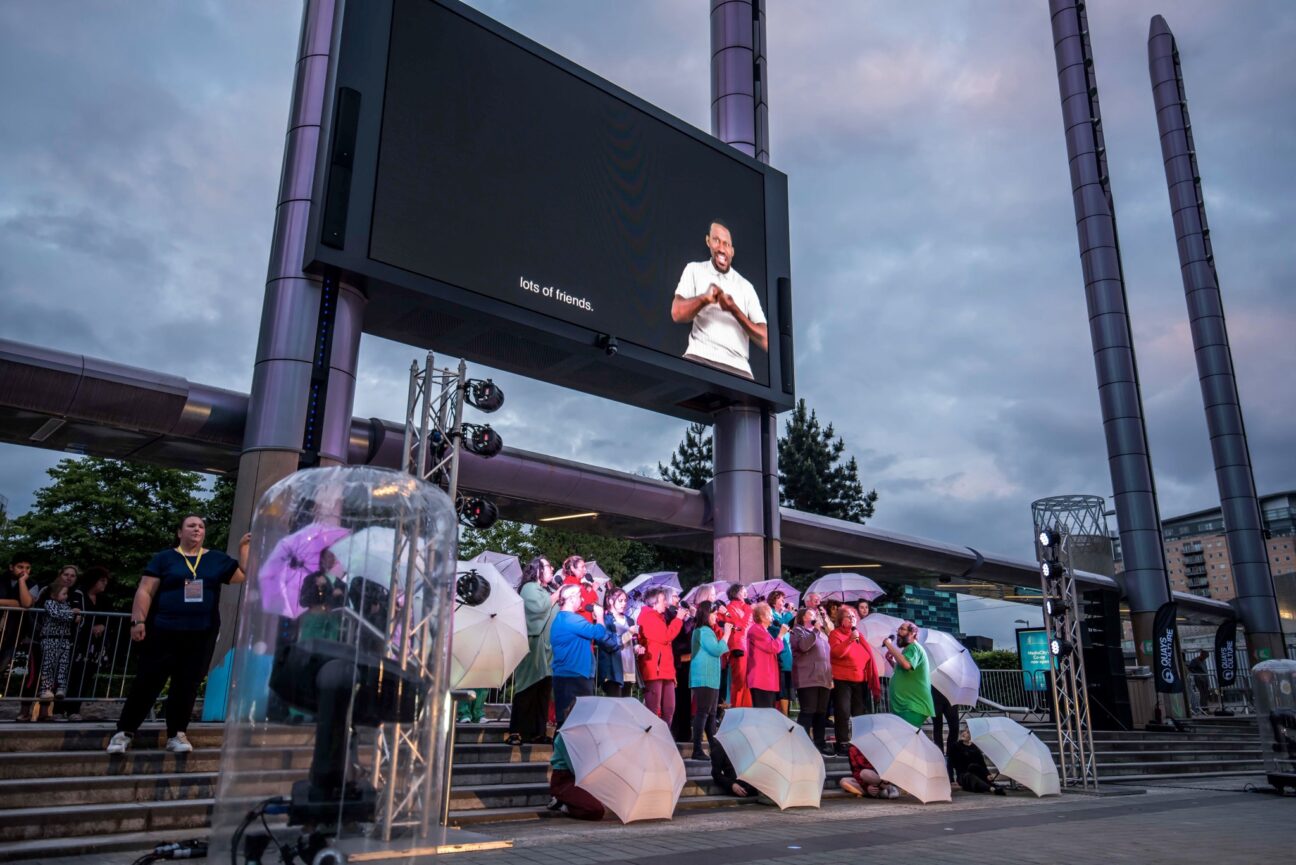 A colourful choir surrounded by umbrellas, hold mics as they sing together directed by a man in a green shirt. Above them is a screen where lyrics are displayed along with a BSL interpreter.