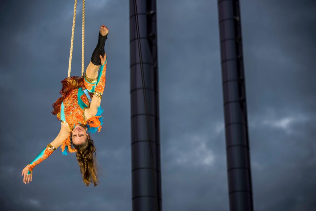 A female aerial performer smiles whilst holding her leg in a split, hanging off of a rope in the air. She is wearing an orange and blue sparkly leotard.