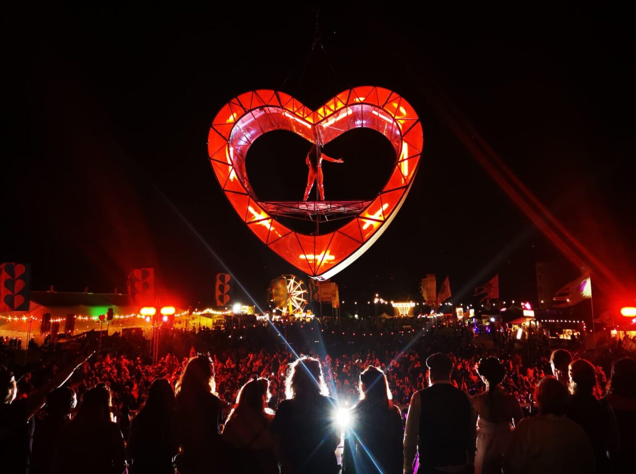 A view from the back of a female singer stood in red metal heart structure above ground with the audience watching from below.