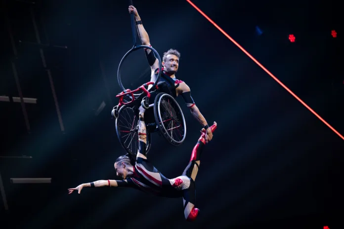 Two aerialists in red and black costumes are suspended in the air against a dark background. Jonny is sitting in a wheelchair and hanging by one hand from a strop. Charlotte, below, holds the bottom of Jonny's wheelchair and strikes a graceful pose supported by Jonny.