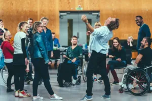 A group of people gather together, in a circle. They are standing or seated. They are smiling. They watch Ted Barnes, who is catching something.