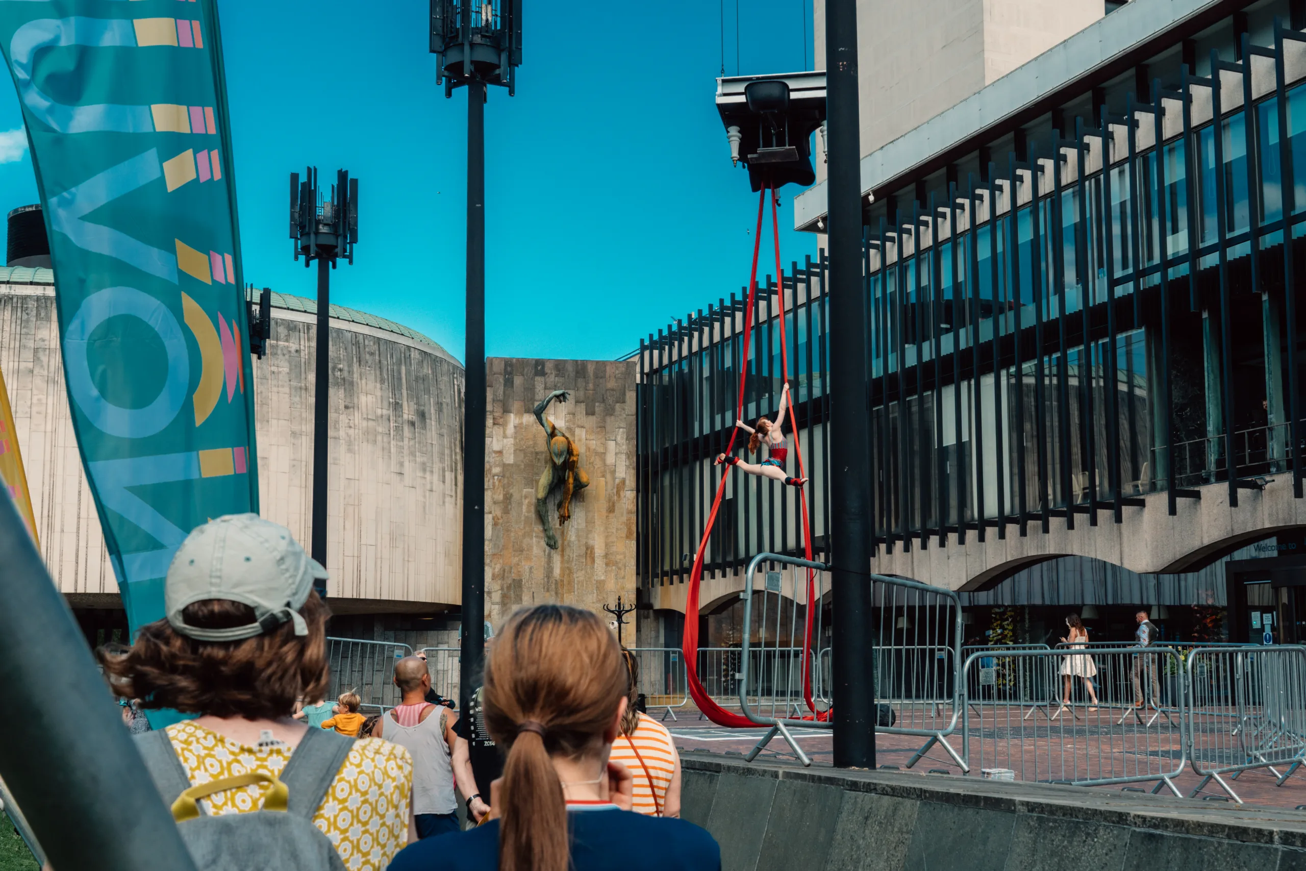 An aerial performer on red silks is suspended beneath a flying piano in an outdoor setting.