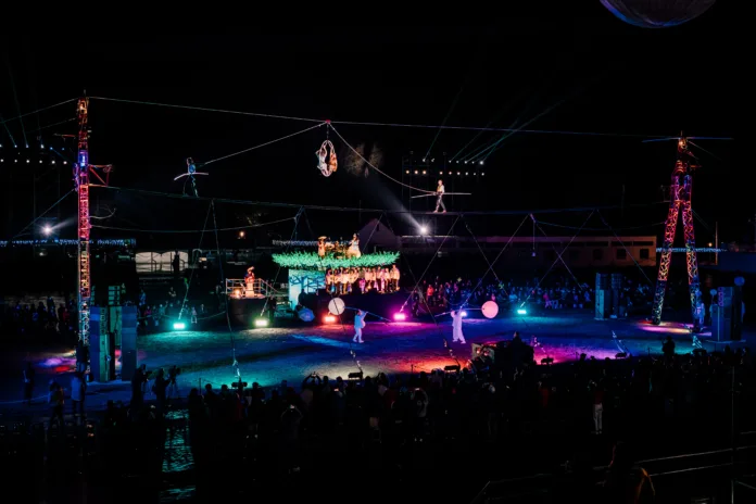 A shot of the high wire, illuminated with lights. Two aerialists dangle and perform from the wire, whilst a dance with illuminated lanterns on sticks happens below