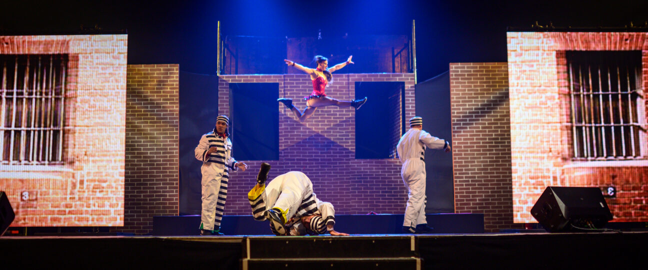 A performer is caught mid leap on a trampoline, jumping over three characters below her